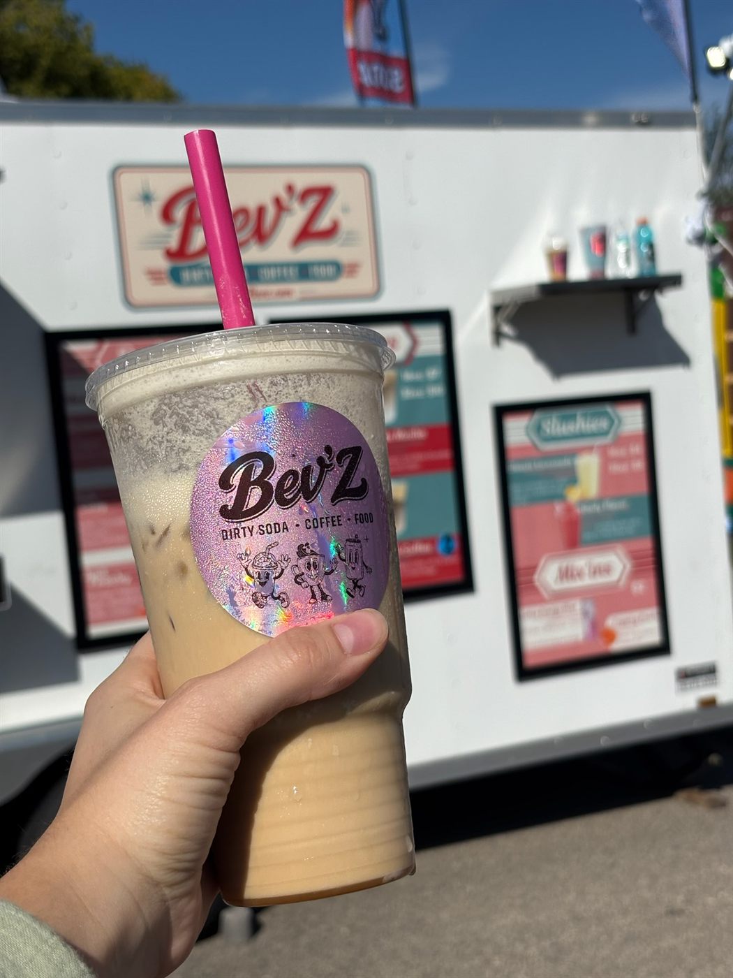 A Bev'Z iced coffee held up in front of the Bev'Z trailer and menu boards